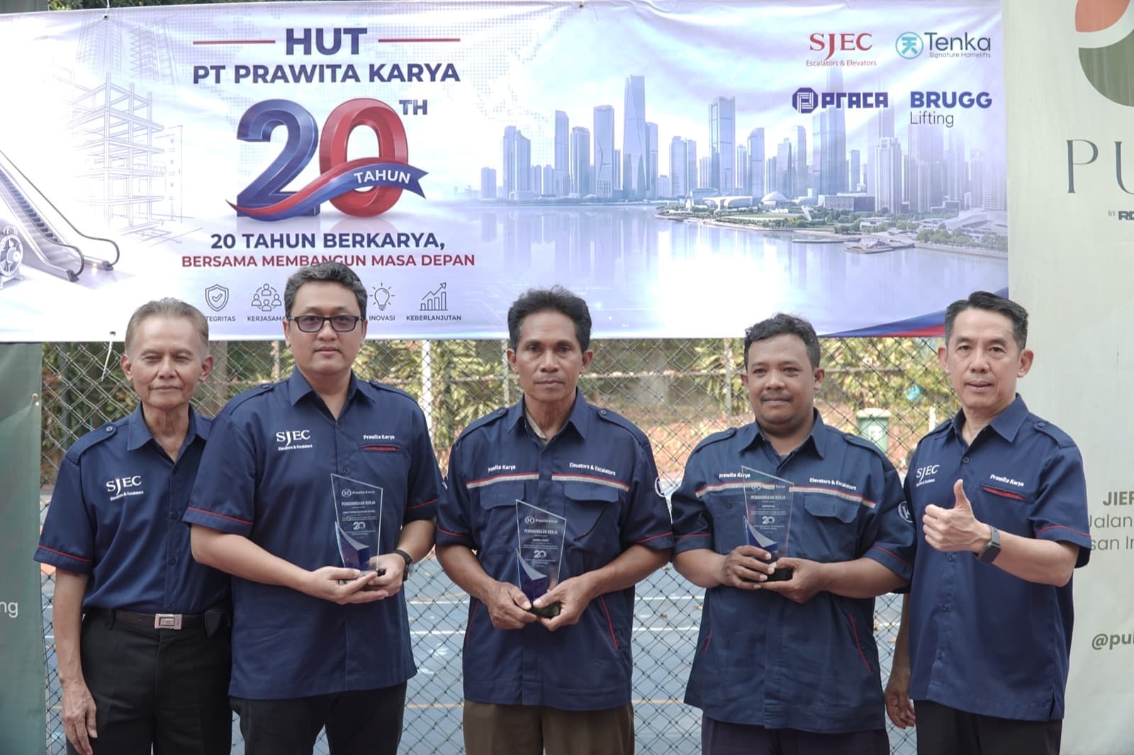 Three Prawita Karya staff holding their twenty-year service awards, with company directors either side, in front of the HUT PT Prawita Karya 20th anniversary banner