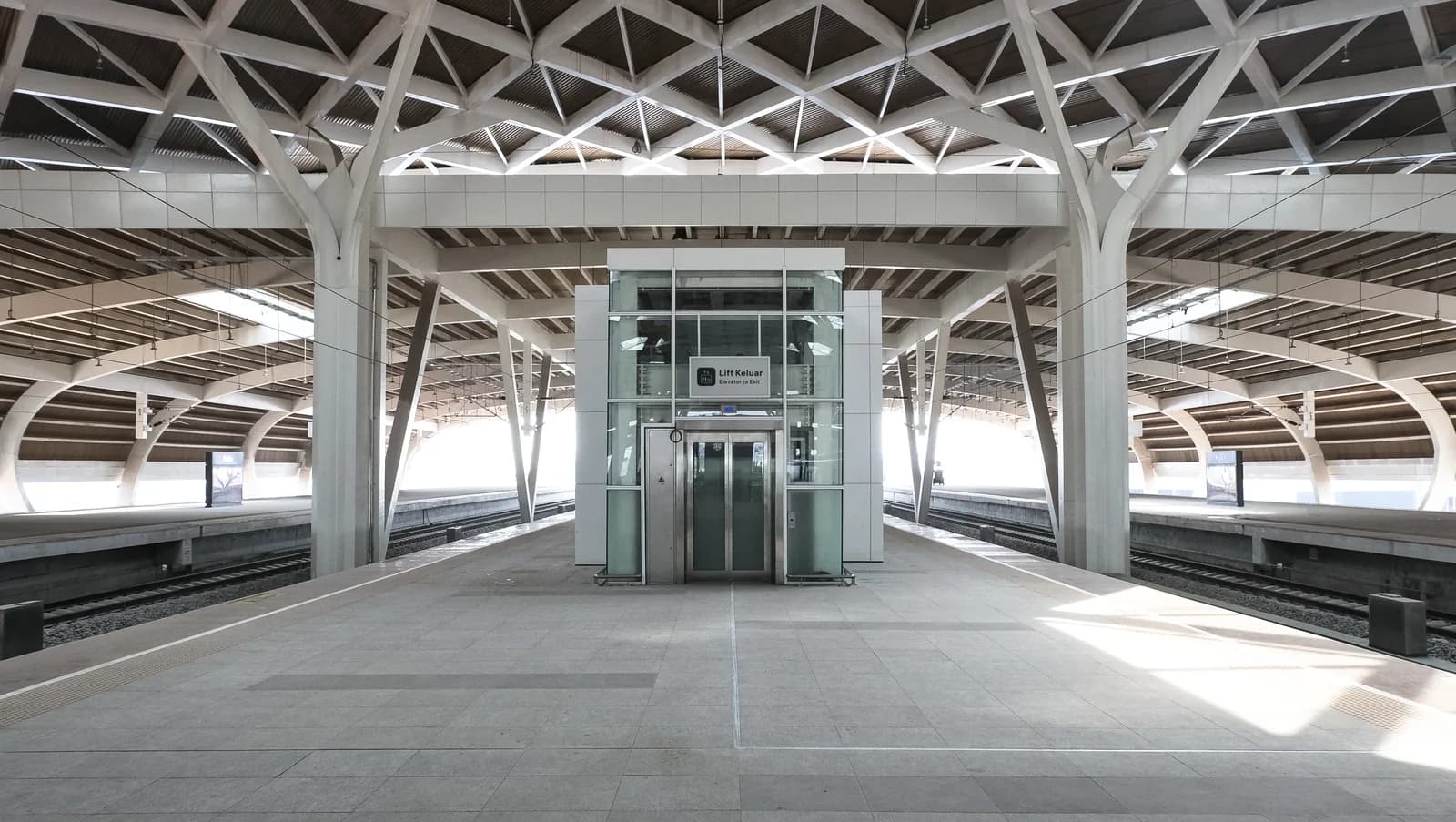 Glass platform elevator signed Lift Keluar / Elevator to Exit standing between the tracks at KCIC Halim high-speed rail station, Jakarta, beneath the station's branching column canopy