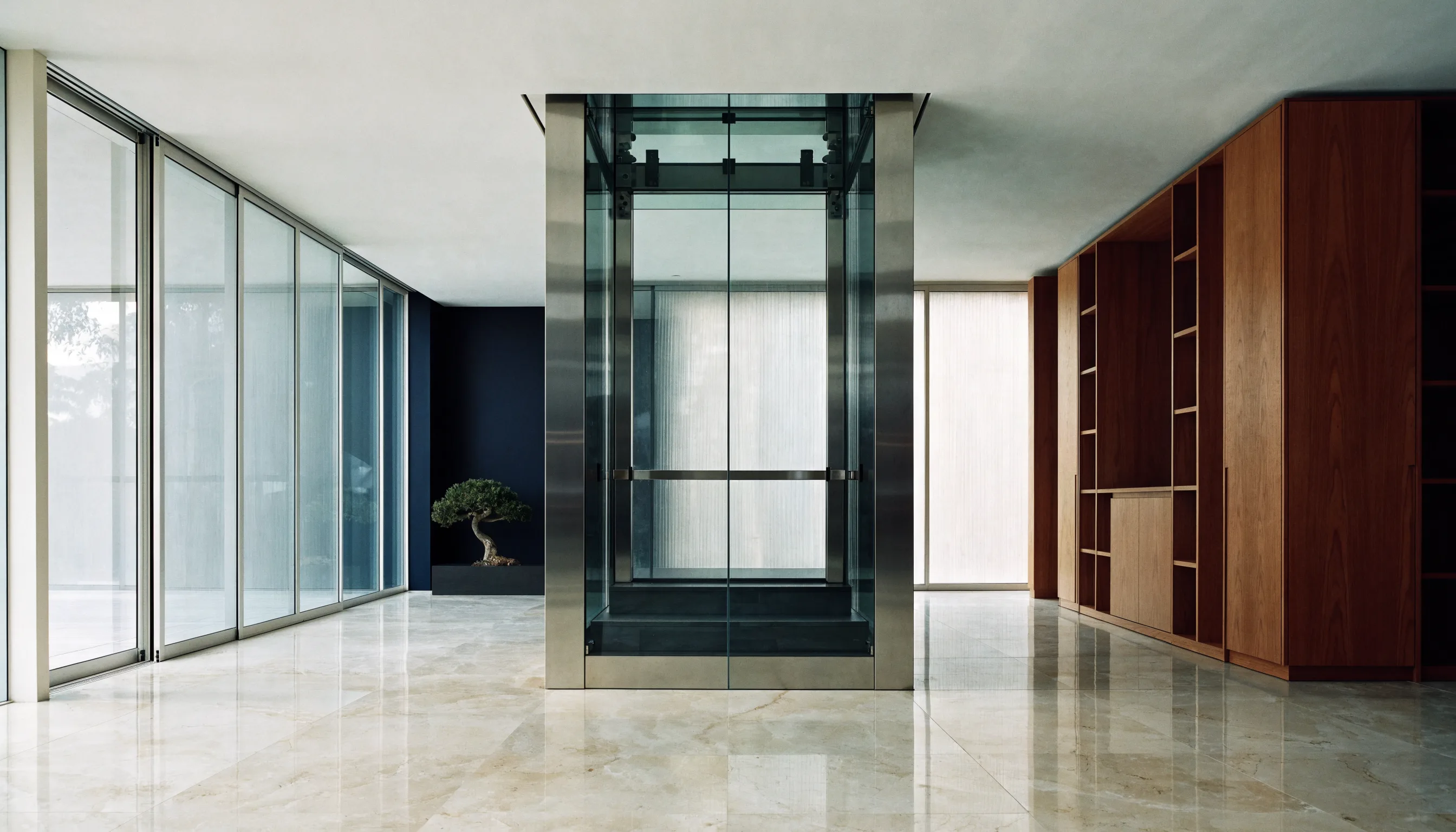 Compact panoramic home lift with a curved glass shaft beside a pale oak stair in a contemporary residence